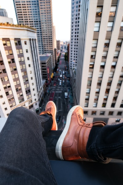 穿着黑色牛仔裤和棕色高帮运动鞋的人坐在屋顶上，可以看到建筑物之间的街道